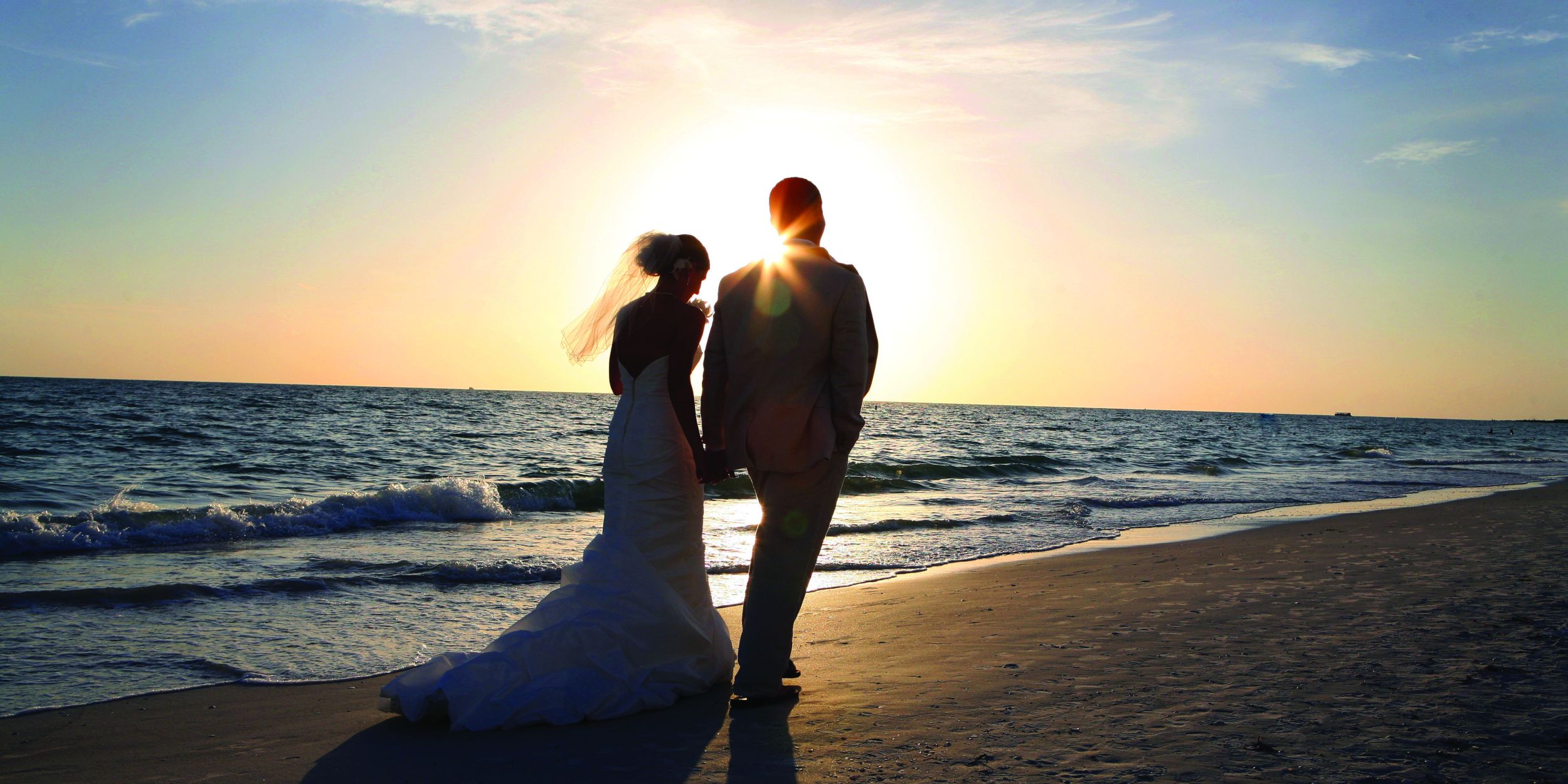 A Man And Woman Standing On A Beach With The Sun Setting Behind Them