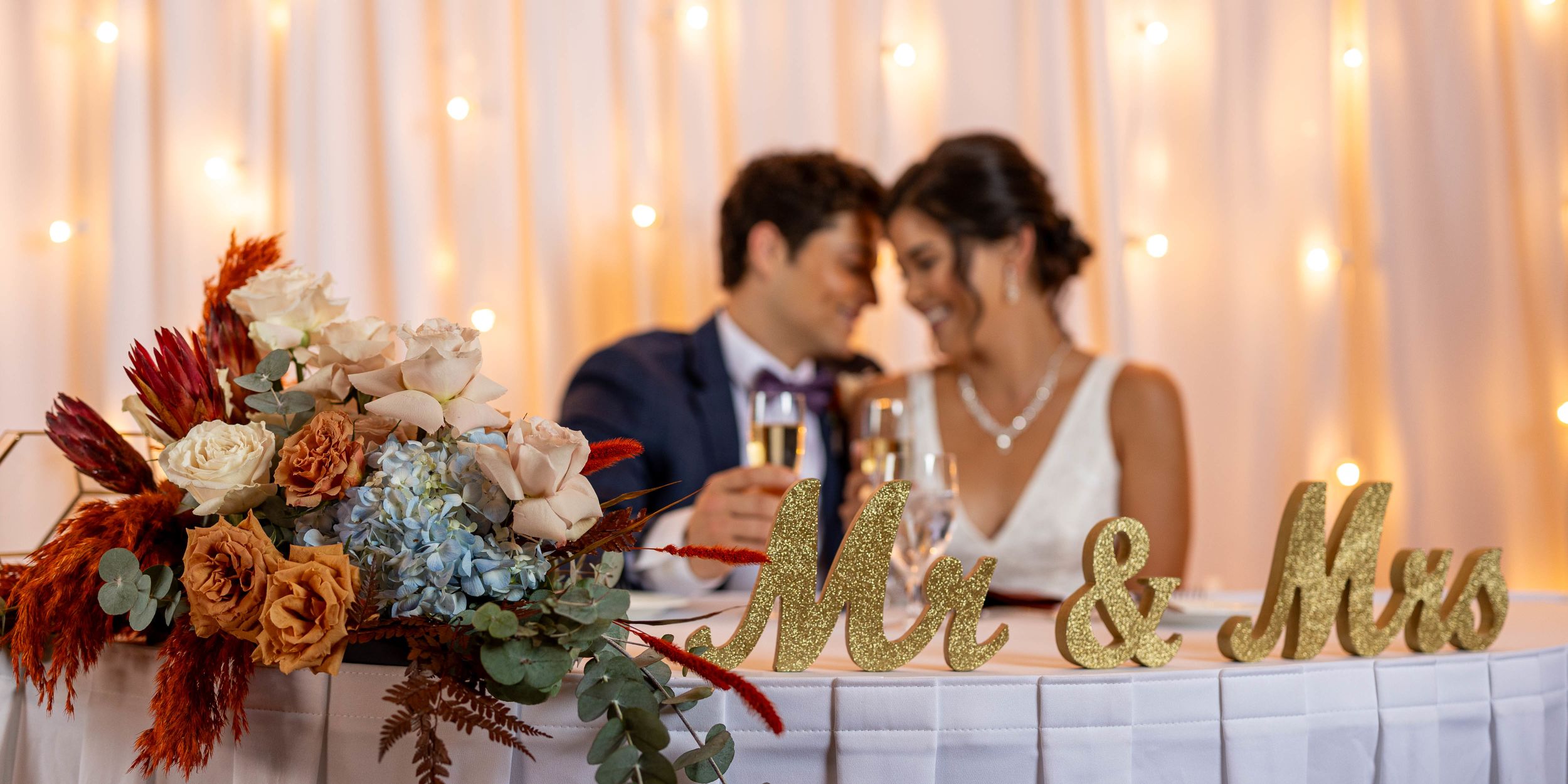 A Bride And Groom Sitting At A Table With Flowers